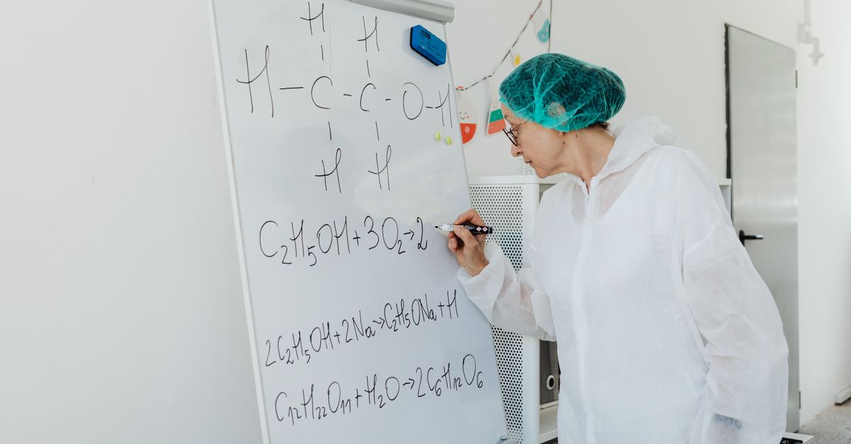 Elderly woman in lab coat writing chemical equations on a whiteboard indoors.
